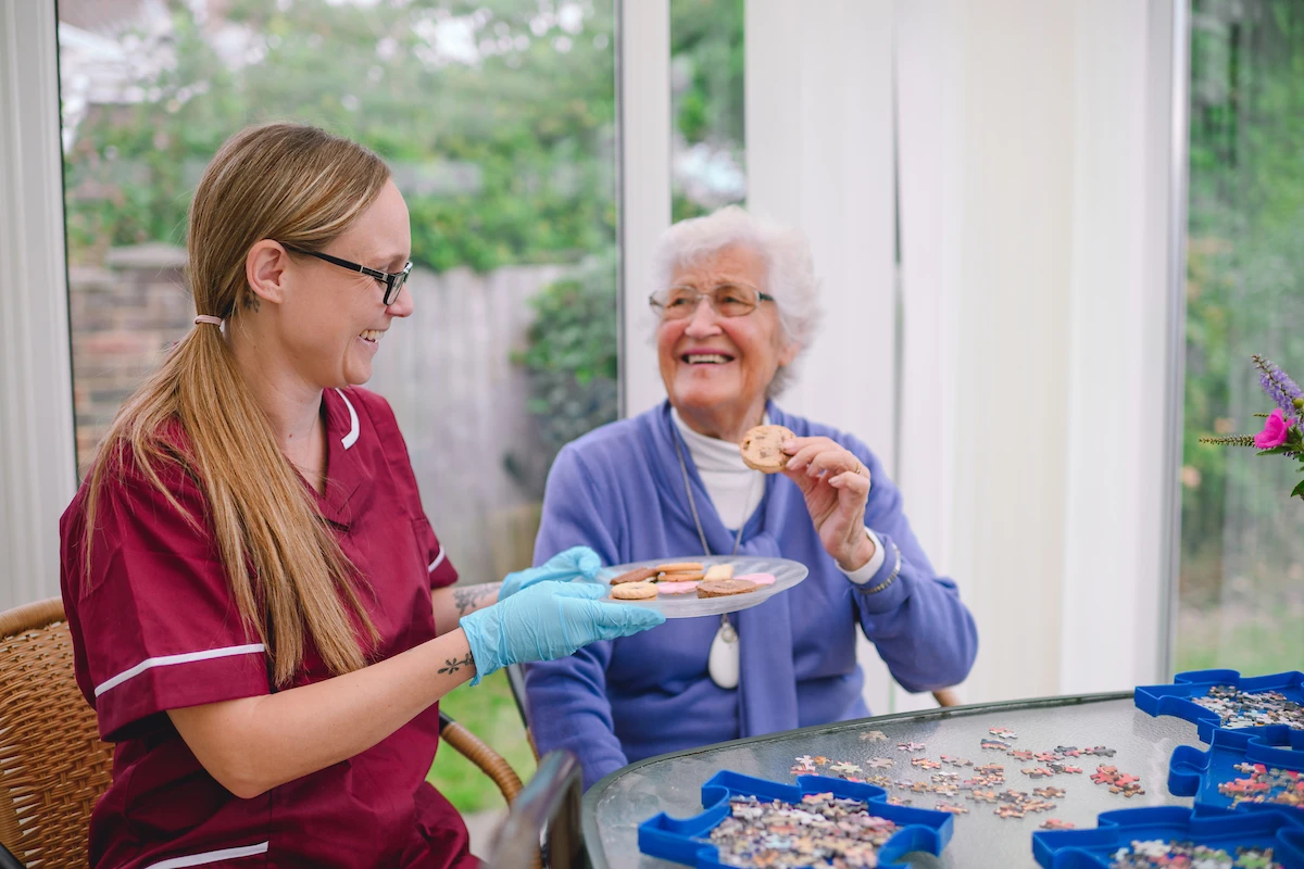 Nestwell home care assistant serving biscuits to an elderly woman during a puzzle activity.